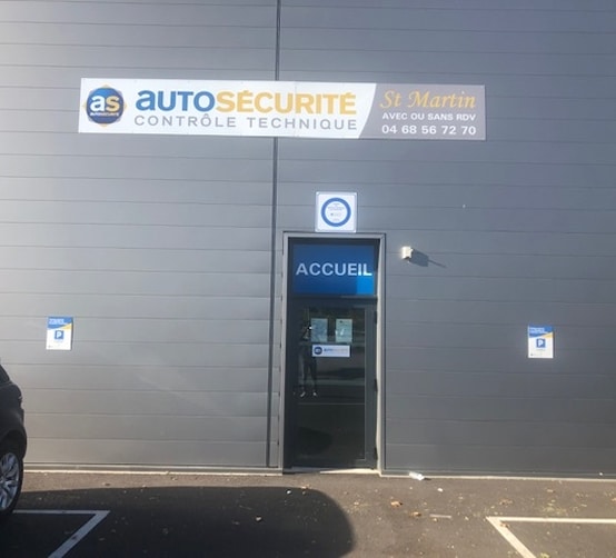 Façade grise d'un centre de contrôle technique AS AutoSécurité St Martin. Le panneau indique "CONTRÔLE TECHNIQUE" et le numéro 04 68 56 72 70. L'entrée est marquée "ACCUEIL".