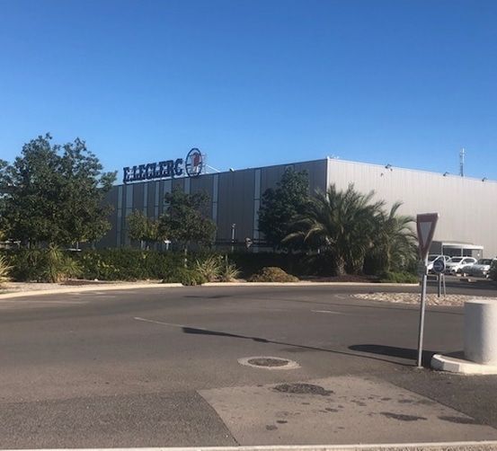 Bâtiment du supermarché E. Leclerc sous un ciel bleu clair, avec une végétation méditerranéenne et un panneau de céder le passage au premier plan.