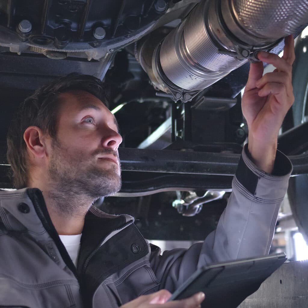 Mécanicien vêtu d'une veste grise examinant et ajustant un collier de serrage sur un tuyau d'échappement flexible de voiture avec une tablette à la main.